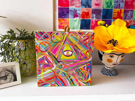 An abstract painting titled All-Seeing Eye from My Store, featuring vibrant geometric shapes and an eye design, sits on a shelf. Its paired with a head vase containing a yellow flower, leafy plant, framed sketch, and backed by a multicolored photo collage.