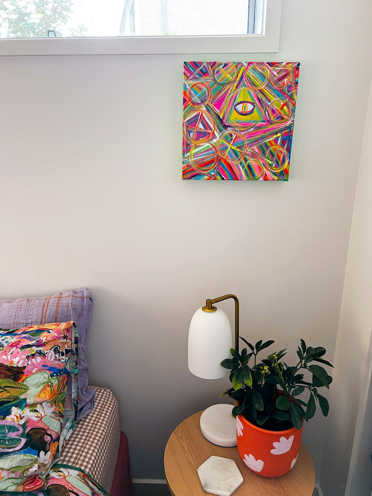 A colorful acrylic painting of geometric shapes and an All-Seeing Eye from My Store adorns the white wall. Below, a small round table holds a potted plant in an orange pot and a white lamp. A patterned bed peeks into view on the left, enhancing the mystical artwork ambiance.