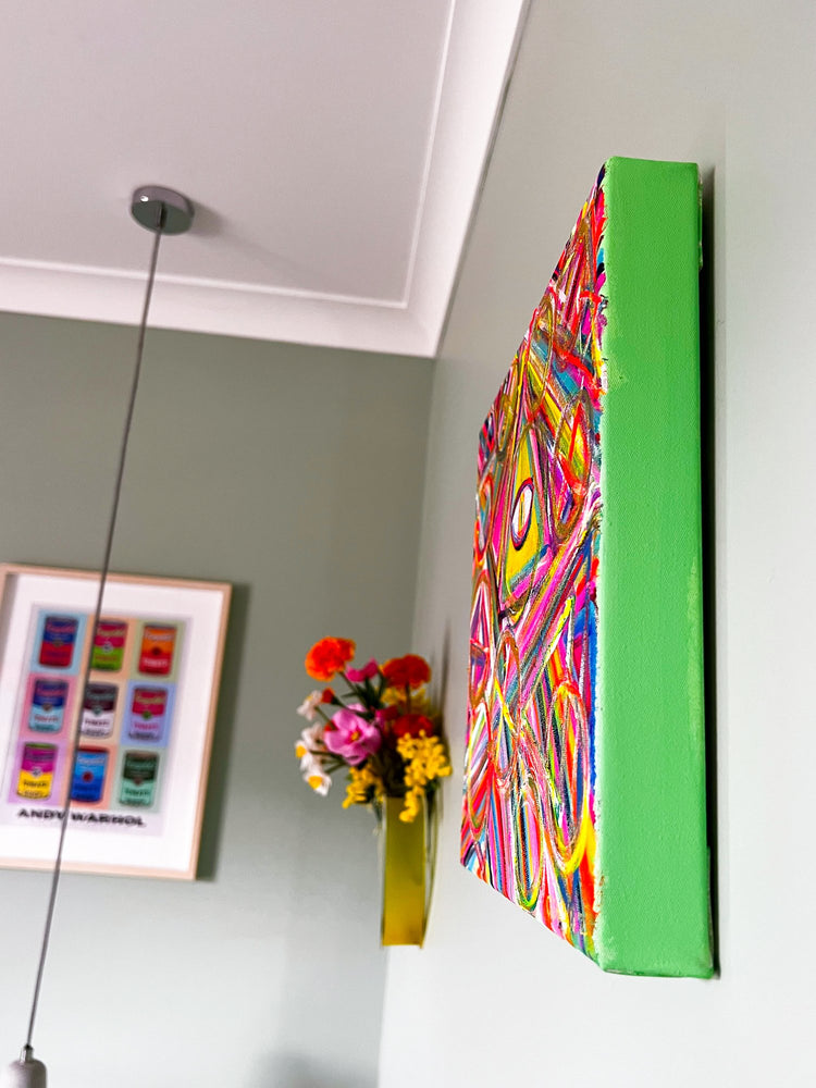A vibrant abstract painting with a green edge is on a light green wall. Below, My Stores All-Seeing Eye Original Painting on canvas stands by a vivid flower bouquet in a small yellow vase, next to framed artwork showcasing multiple color variations.