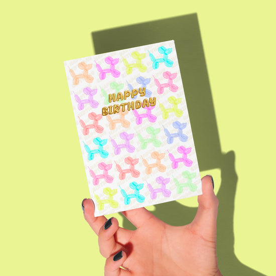 A hand displays My Stores Balloon Dog Birthday Card, featuring colorful balloon dogs on a light background. HAPPY BIRTHDAY shines in gold at the center, ideal for celebrating. The backdrop is a cheerful light green.