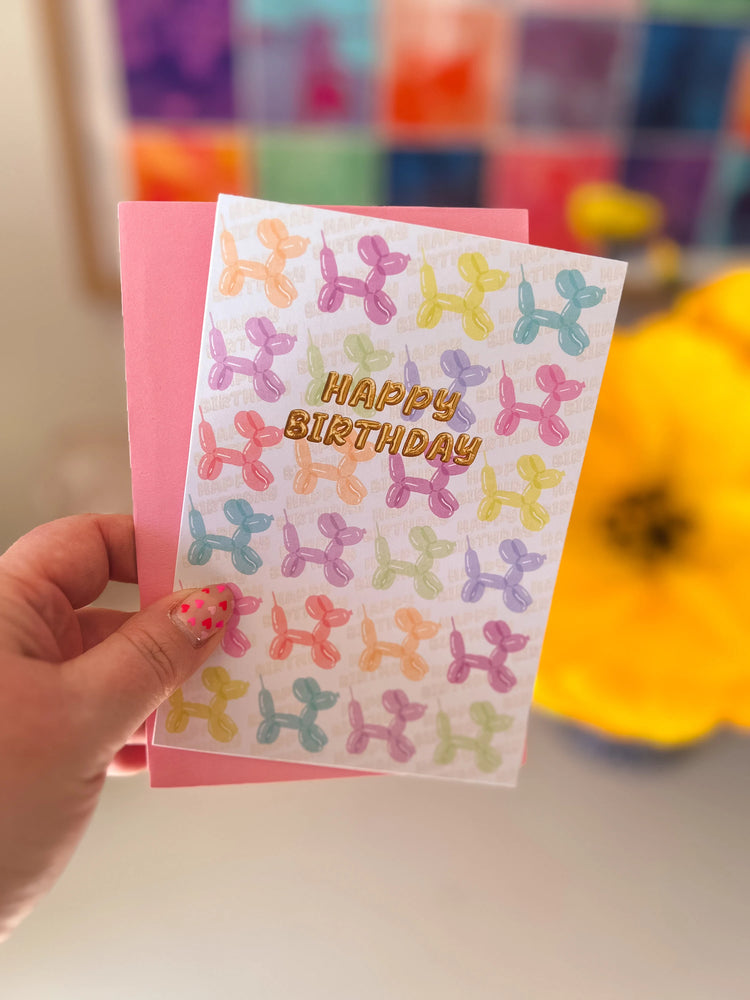 A hand displays My Stores Balloon Dog Birthday Card, featuring balloon animal designs and HAPPY BIRTHDAY in gold. Polka dot nail art adds charm, while a pink envelope and blurred yellow flower form a delightful backdrop for celebrating a special day.