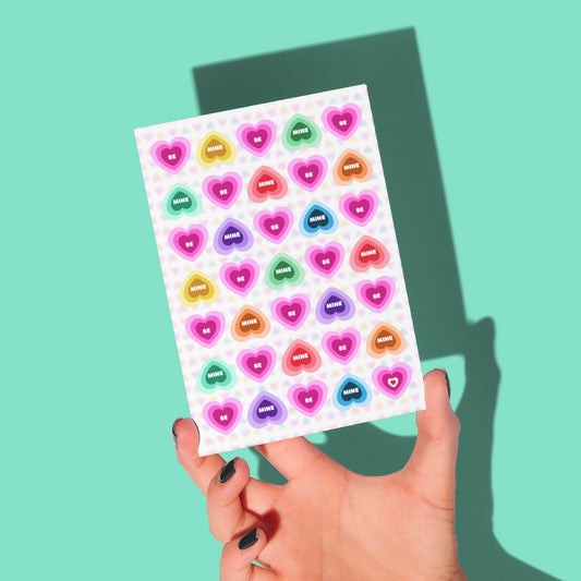 A hand displays the Be Mine Valentines Card against a teal backdrop. The card features rainbow candy hearts in a grid.