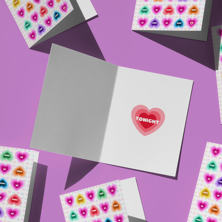 A variety of greeting cards lie on a purple surface. At the centre, an open Be Mine Valentines card from My Favourite Colour is Rainbow displays TONIGHT inside a pink heart. Surrounding it, closed cards feature vibrant patterns similar to rainbow candy hearts.