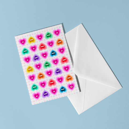 The Be Mine Valentines Card by My Store features rainbow candy hearts with words like happy and smile, set against a pastel blue background, accompanied by a white envelope.