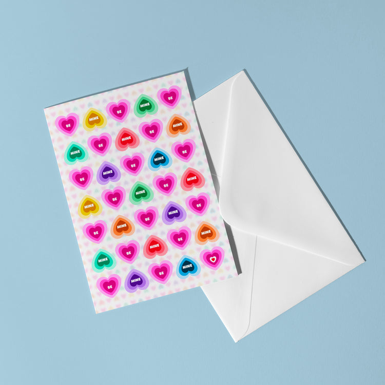 The Be Mine Valentines Card by My Store features rainbow candy hearts with words like happy and smile, set against a pastel blue background, accompanied by a white envelope.