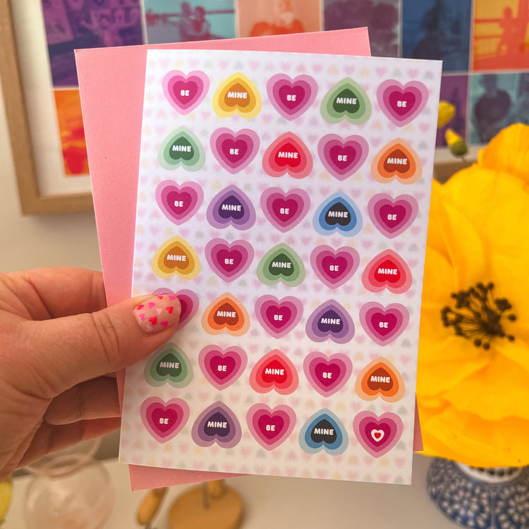 A hand holds a sheet of heart-shaped stickers with BE MINE in various colors, similar to rainbow candy hearts, next to a My Store Be Mine Valentines Day card. A bright yellow flower blooms nearby, with a colorful board enhancing the walls charm.