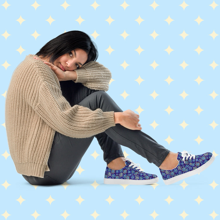 A person with chin-length dark hair sits on the floor, resting their head on their knee. They wear a beige sweater, dark pants, and Bloomscape Canvas Sneakers by My Favourite Colour is Rainbow, featuring a blue floral design.