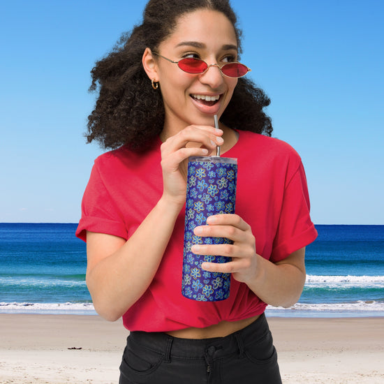Wearing a red shirt and sunglasses, a woman smiles while sipping from her My Favourite Colour is Rainbow Bloomscape Stainless Steel Tumbler with a metal straw. She stands before a sunny beach, enjoying the blue waves and clear sky.