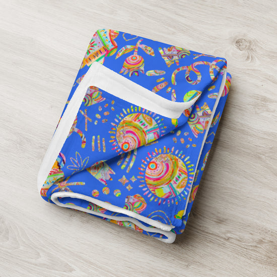 The Blue Summer Sorbet Throw Blanket by My Favourite Colour is Rainbow features a vibrant abstract design with swirling patterns and geometric shapes. Primarily in blue hues, it adds a splash of color to any room while showcasing its detailed print and cosy, soft texture on a light wooden surface.