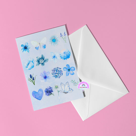 The Blue Watercolour Greeting Card by My Store, featuring butterfly magic and blue flowers, rests next to a white envelope on a pink background. A rainbow sticker creatively seals the envelope, adding a whimsical touch.