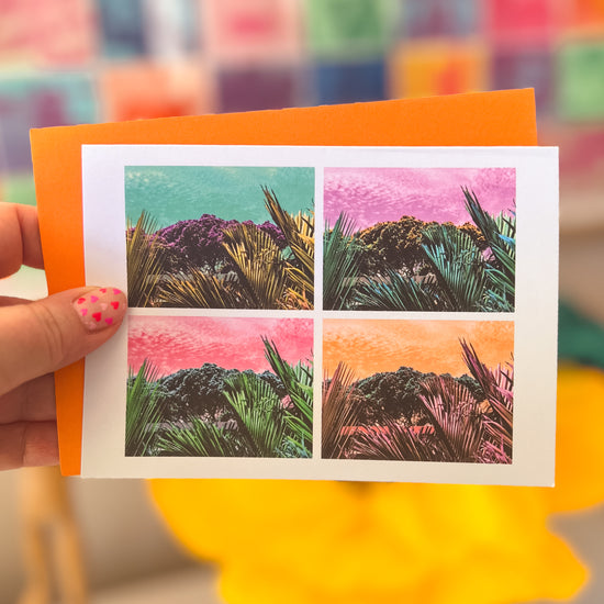 A hand displays My Stores Pohutukawa Pop Art greeting card (Version 3) featuring palm trees and hills in vibrant panels, reminiscent of New Zealands charm. It rests on an orange envelope against a blurred square backdrop.