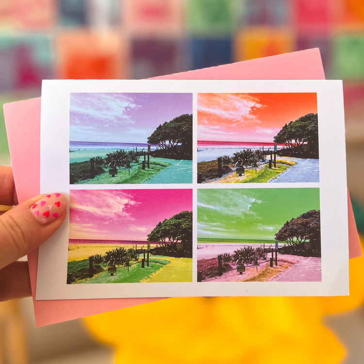 A hand with pink heart-patterned nails holds the My Store Pohutukawa Pop Art greeting card (Version 2), showcasing a New Zealand beach scene in purple, orange, pink, and green. The vibrant postcard is set against a blurred, colorful background.