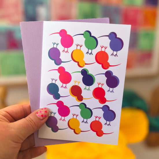 A person holds a Technicolour Kiwi greeting card by My Store, featuring colorful kiwis and bird pairs in red, orange, yellow, green, blue, and purple. A lavender envelope is tucked behind it against a softly blurred background.