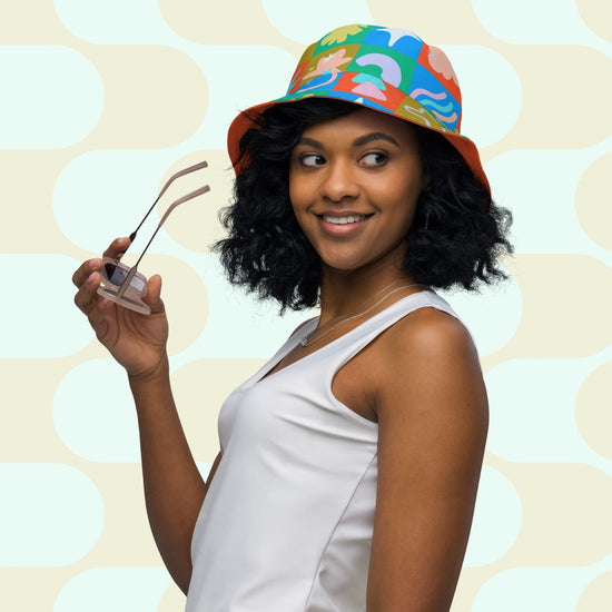 A woman with curly hair smiles while holding sunglasses, wearing a white tank top and the Colour Block Reversible Bucket Hat by My Favourite Colour is Rainbow.