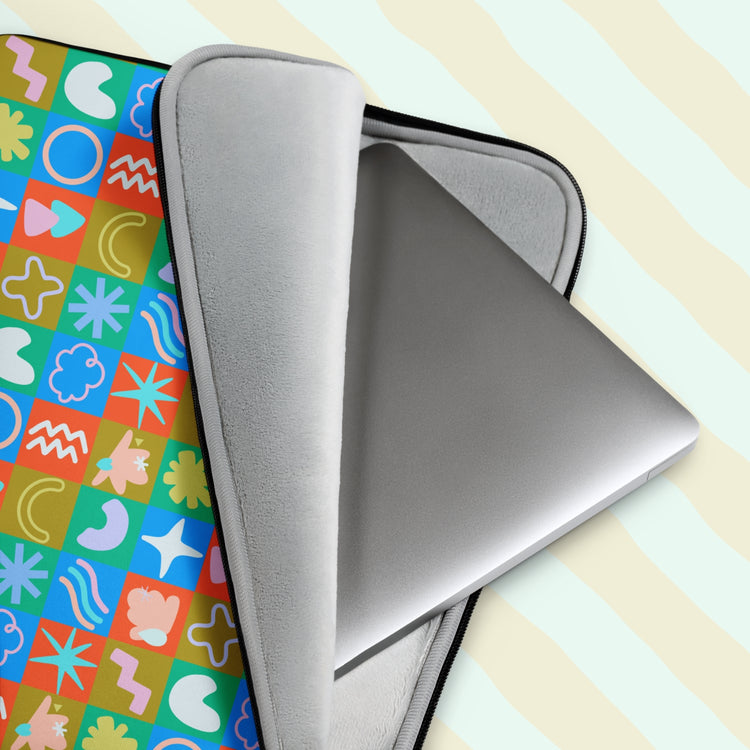 A laptop partially slides out of the Colour Block Laptop Sleeve by My Favourite Colour is Rainbow, featuring a grid of abstract shapes in colours with a vintage vibe.  It has a grey padded interior to keep your laptop safe from scratches.  The print is on only one side of the laptop sleeve, the other side is black. It is shown on a pastel background.