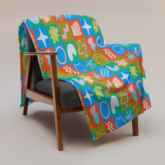 A mid-century modern wooden chair with gray cushions is enhanced by My Favourite Colour is Rainbows Colour Block Throw Blanket, featuring stars, hearts, and swirls in red, blue, green, and orange on a neutral beige background.