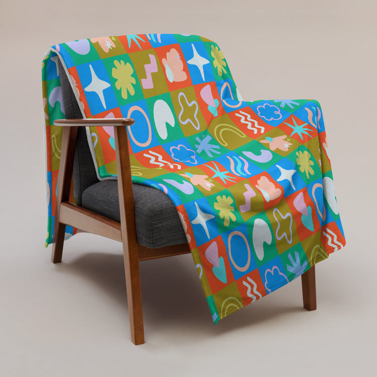 A mid-century modern wooden chair with gray cushions is enhanced by My Favourite Colour is Rainbows Colour Block Throw Blanket, featuring stars, hearts, and swirls in red, blue, green, and orange on a neutral beige background.