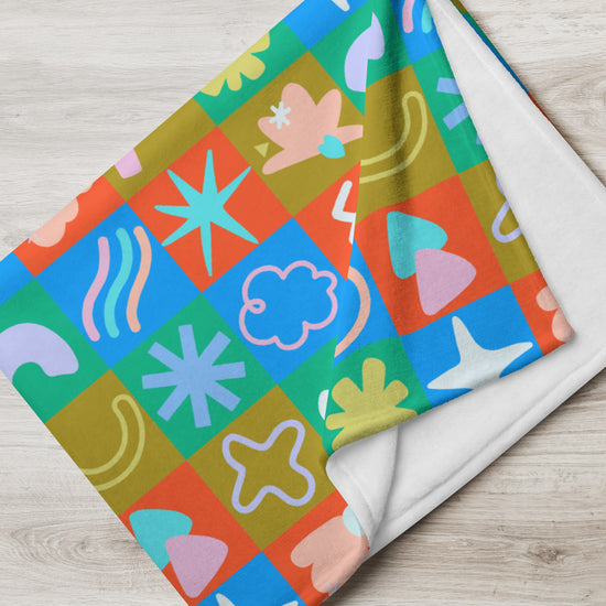 The Colour Block Throw Blanket by My Favourite Colour is Rainbow showcases a lively design with abstract grid patterns, featuring stars, wavy lines, clouds, and organic shapes in blue, orange, green, and pink on its soft fabric atop a wooden surface.
