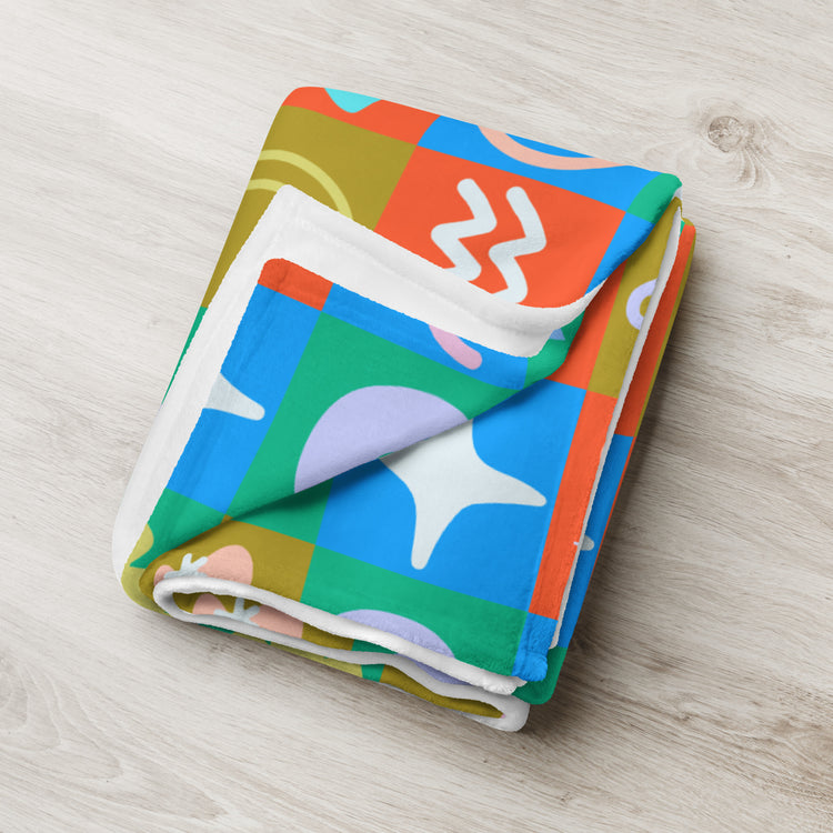 The Colour Block Throw Blanket by My Favourite Colour is Rainbow lies folded on a light wooden surface, showcasing abstract patterns with vibrant squares in blue, orange, green, and white. Its whimsical design adds a playful modern touch while the soft fabric ensures cozy comfort in any room.