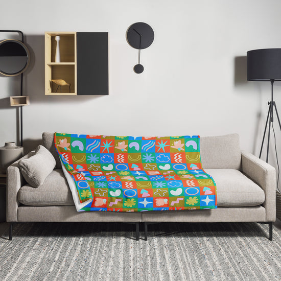 A modern living room with a gray sofa features a vibrant design, with the My Favourite Colour is Rainbow Colour Block Throw Blanket, crafted from soft fabric and geometric patterns, draped over it. The minimalist wall has a wooden shelf, round clock, and black floor lamp on the right.