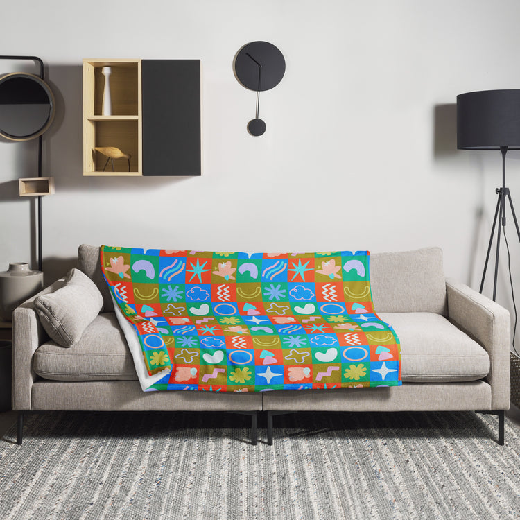 A modern living room with a gray sofa features a vibrant design, with the My Favourite Colour is Rainbow Colour Block Throw Blanket, crafted from soft fabric and geometric patterns, draped over it. The minimalist wall has a wooden shelf, round clock, and black floor lamp on the right.