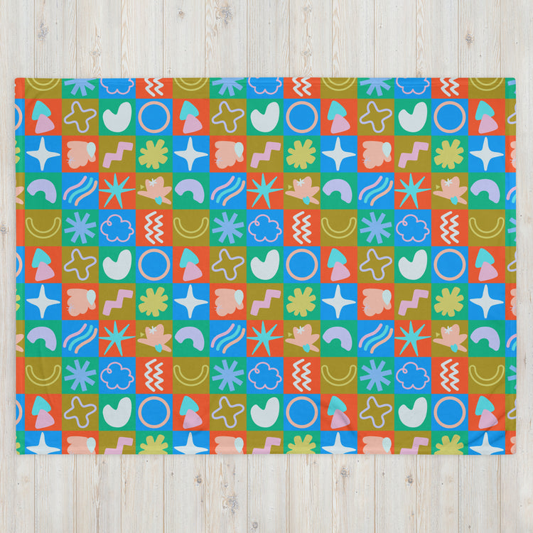 The Colour Block Throw Blanket by My Favourite Colour is Rainbow features a lively design with colorful geometric shapes—circles, stars, squiggles, and abstract forms in orange, red, blue, green, and pink. Its soft fabric pops against a light wooden backdrop.