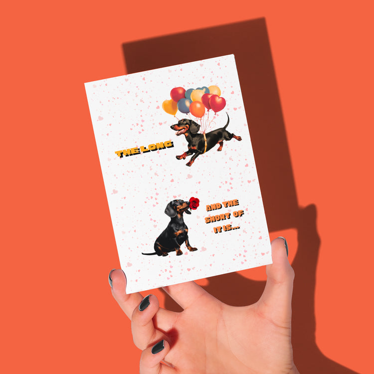 A hand holds the "Perfect Match" Dachshund Valentines Card against an orange backdrop. It features cartoon sausage dogs—one with balloons, the other with a rose—and text" The long and And the short of it..."