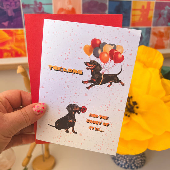 A hand holds a Perfect Match Dachshund Valentines card from My Store, showcasing two dachshunds: one lifted by balloons and the other with a red rose. The card reads, The long and the short of it is... against a backdrop of colorful artwork, featuring a yellow flower—ideal for expressing love.