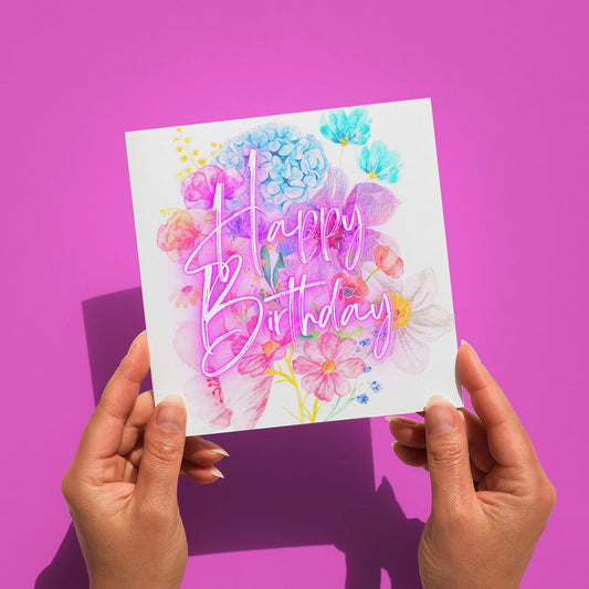 Hands holding the Floral Happy Birthday Card by My Store against a pink background. The square card features vibrant floral designs and Happy Birthday in elegant pink cursive lettering.