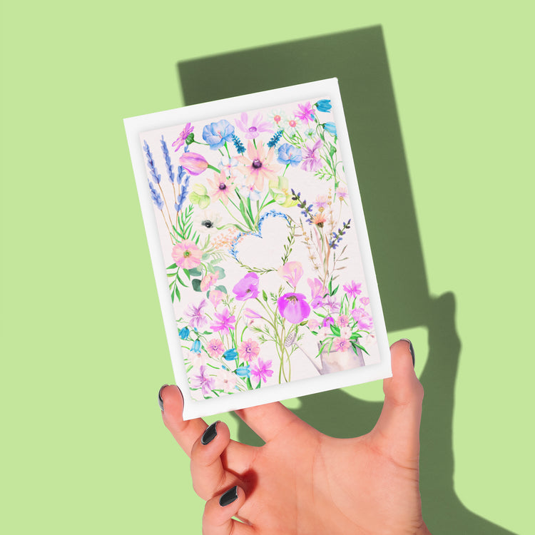 A hand with dark nail polish holds the My Store Floral Watercolour Love Greeting Card, featuring pink, purple, and blue flowers around a heart-shaped cutout. It casts a shadow on the light green background.