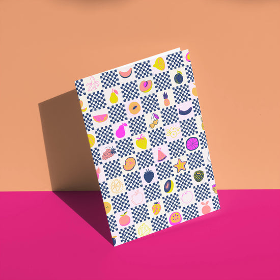 A Fruit Salad Greeting Card from My Store, featuring a colorful pineapple and geometric pattern, is displayed upright on a tan and pink surface. Shadows cast to the left enhance the vibrant design.