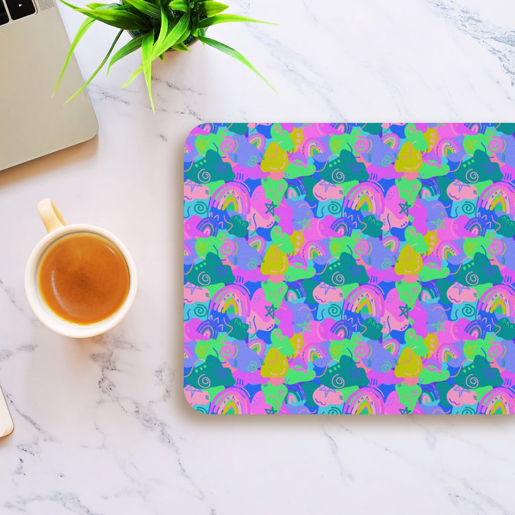 The Funfetti Mouse Pad by My Favourite Colour is Rainbow features abstract shapes in pink, green, and blue on a white marble desk. A coffee cup sits on the left and a silver laptop is partially visible, with a small green plant in the top left corner.