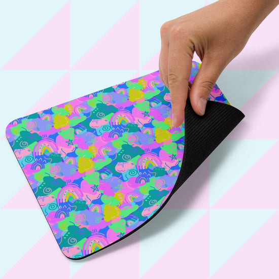 A hand holds the Funfetti Mouse Pad by My Favourite Colour is Rainbow, adorned with abstract shapes in vibrant pink, blue, green, and yellow. The background boasts a pastel pink and blue geometric design.