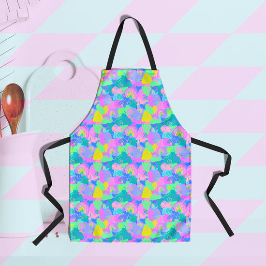 Meet the Funfetti Apron by My Favourite Colour is Rainbow, adorned with a riot of pink, yellow, and blue abstract shapes and rainbows. It has black straps.  A chopping board, utensil holder and wooden spoon are in the background.
