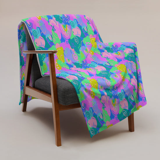 A wooden armchair with a gray cushion showcases a Funfetti Throw Blanket by My Favourite Colour is Rainbow, adorned with vibrant abstract patterns in pink, green, blue, and yellow, creating a striking contrast against the plain beige background.