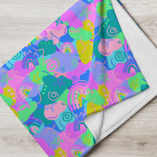 The My Favourite Colour is Rainbows Funfetti Throw Blanket boasts a vibrant design with brightly colored abstract shapes like spirals, stars, and rainbows in pink, blue, green, and yellow. The cozy blanket is partially folded on a light wooden surface.