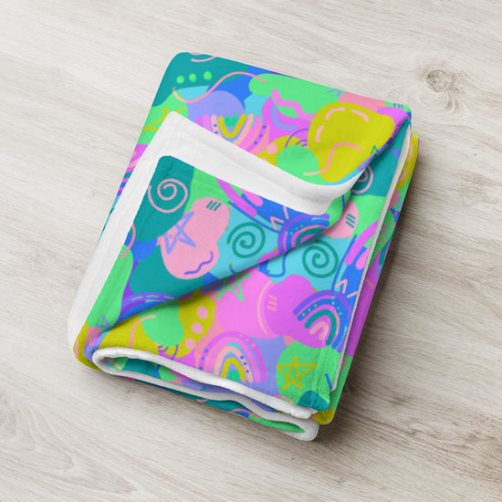 The Funfetti Throw Blanket by My Favourite Colour is Rainbow features an abstract pattern of swirls, stars, and arcs in bright pink, green, blue, and yellow. Elegant on a light wooden surface, it brings vibrant color to any space.