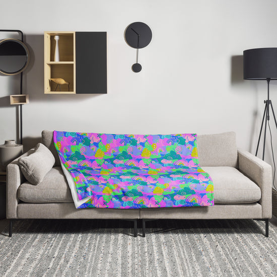 The living room features a light gray sofa with a vibrant, cosy design. Draped over it is the My Favourite Colour is Rainbow Funfetti Throw Blanket with floral pattern. A circular black wall clock hangs above, with a black standing lamp to the right and shelves of decor items to the left.