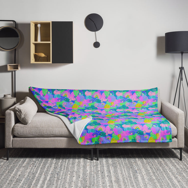 A beige sofa is adorned with a vibrant Funfetti Throw Blanket by My Favourite Colour is Rainbow in pink, purple, green, and blue. The room includes a black floor lamp, circular black wall clock, a decorative wall shelf, and a cozy gray textured rug.