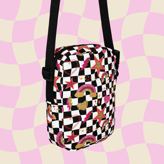 The Galaxy Mini Crossbody Bag by My Favourite Colour is Rainbow features a black and white checkered pattern with gold, pink and red rainbows, stars, and hearts. It is made of canvas-type material, has internal and external pockets and a black strap with a buckle on one side that can be extended to crossbody length.