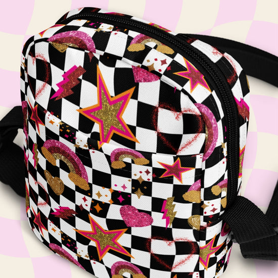 The Galaxy Mini Crossbody Bag by My Favourite Colour is Rainbow features a black and white checkered pattern with glittery gold, pink and red rainbows, stars, and hearts.  It is made of canvas-type material, has internal and external pockets and a black strap with a buckle on one side that can be extended to crossbody length.