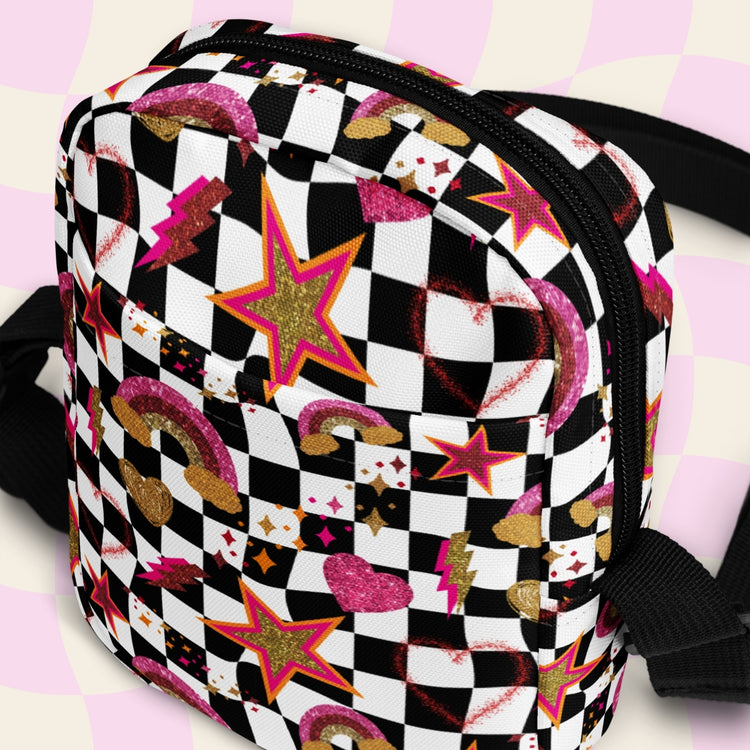 The Galaxy Mini Crossbody Bag by My Favourite Colour is Rainbow features a black and white checkered pattern with glittery gold, pink and red rainbows, stars, and hearts.  It is made of canvas-type material, has internal and external pockets and a black strap with a buckle on one side that can be extended to crossbody length.