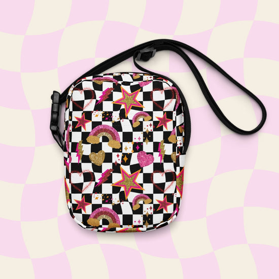 The Galaxy Mini Crossbody Bag by My Favourite Colour is Rainbow is a compact, durable crossbody featuring a black and white checkered pattern with glittery gold, pink and red rainbows, stars, and hearts. It is made of canvas-type material, has internal and external pockets and a black strap with a buckle on one side that can be extended to crossbody length.