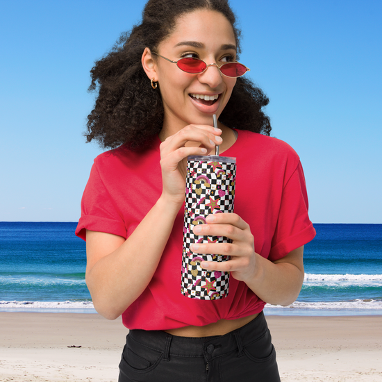 A woman with curly hair and red sunglasses smiles while sipping from a Galaxy Stainless Steel Tumbler by My Favourite Colour is Rainbow. Shes in a red T-shirt, standing on a beach against a vibrant blue sky and ocean backdrop.