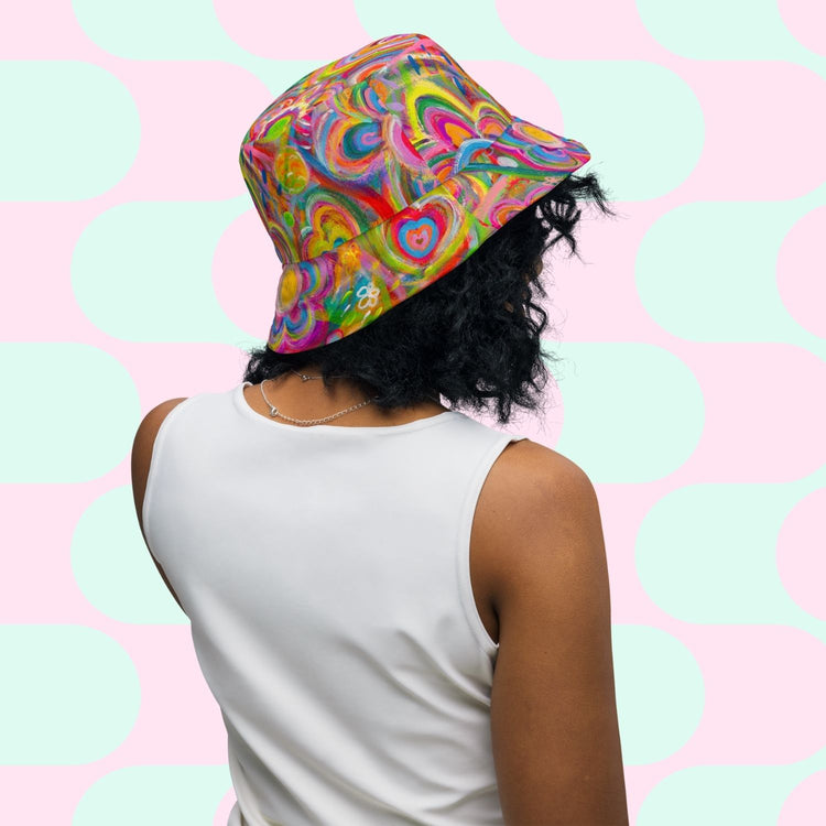 Against a pastel pink and light turquoise geometric background, a person with wavy black hair wears My Favourite Colour is Rainbows Geometric Rainbow/Love in Neon Reversible Bucket Hat and a sleeveless white top, embodying the whimsical allure of a geometric rainbow.