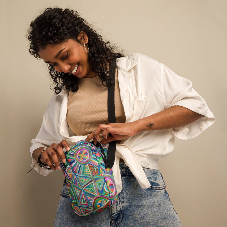 A smiling woman with curly hair wears a tan top and white shirt with light blue jeans, opening the Geometric Rainbow Mini Crossbody Bag by My Favourite Colour is Rainbow, featuring adjustable black straps.