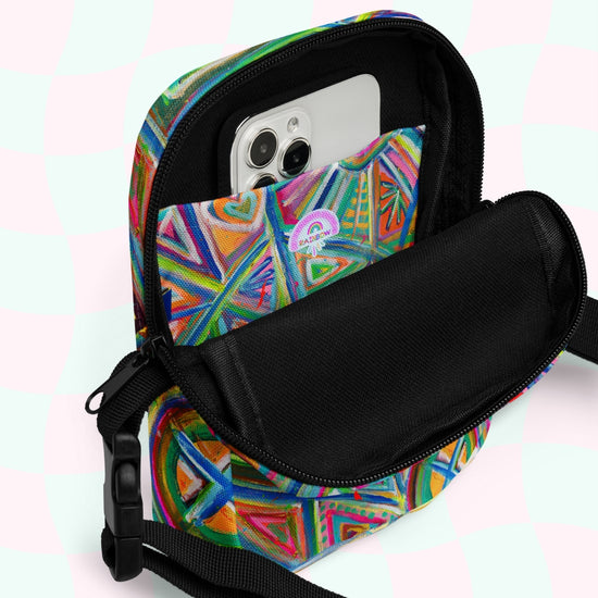 The Geometric Rainbow Mini Crossbody Bag by My Favourite Colour is Rainbow features an adjustable strap and vibrant geometric rainbow print over a pastel checkered background, with an open front pocket partially revealing a smartphone.