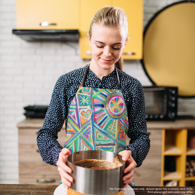 A person in a polka dot shirt and a Geometric Rainbow Apron by My Favourite Colour is Rainbow holds a round cake mold on a wooden countertop. The kitchen, with yellow cabinets and a brick backsplash, sets the perfect backdrop, while appliances are neatly arranged in the background.  The apron features vivid geometric designs in blue, pink, and green