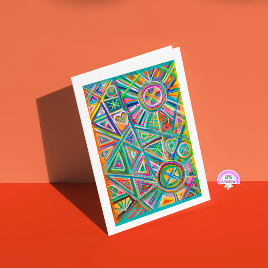 The Geometric Rainbow Greeting Card by My Store features colorful geometric patterns of triangles, circles, and hearts in vivid blues, pinks, and oranges. Its displayed on an orange surface against a matching background with a small geometric rainbow sticker.