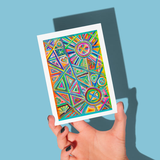 A hand with dark nail polish holds a Geometric Rainbow Greeting Card by My Store against a light blue background, featuring triangles, circles, hearts, and arrows in lively shades of green, orange, pink, and blue. A shadow of the card is cast on the wall.